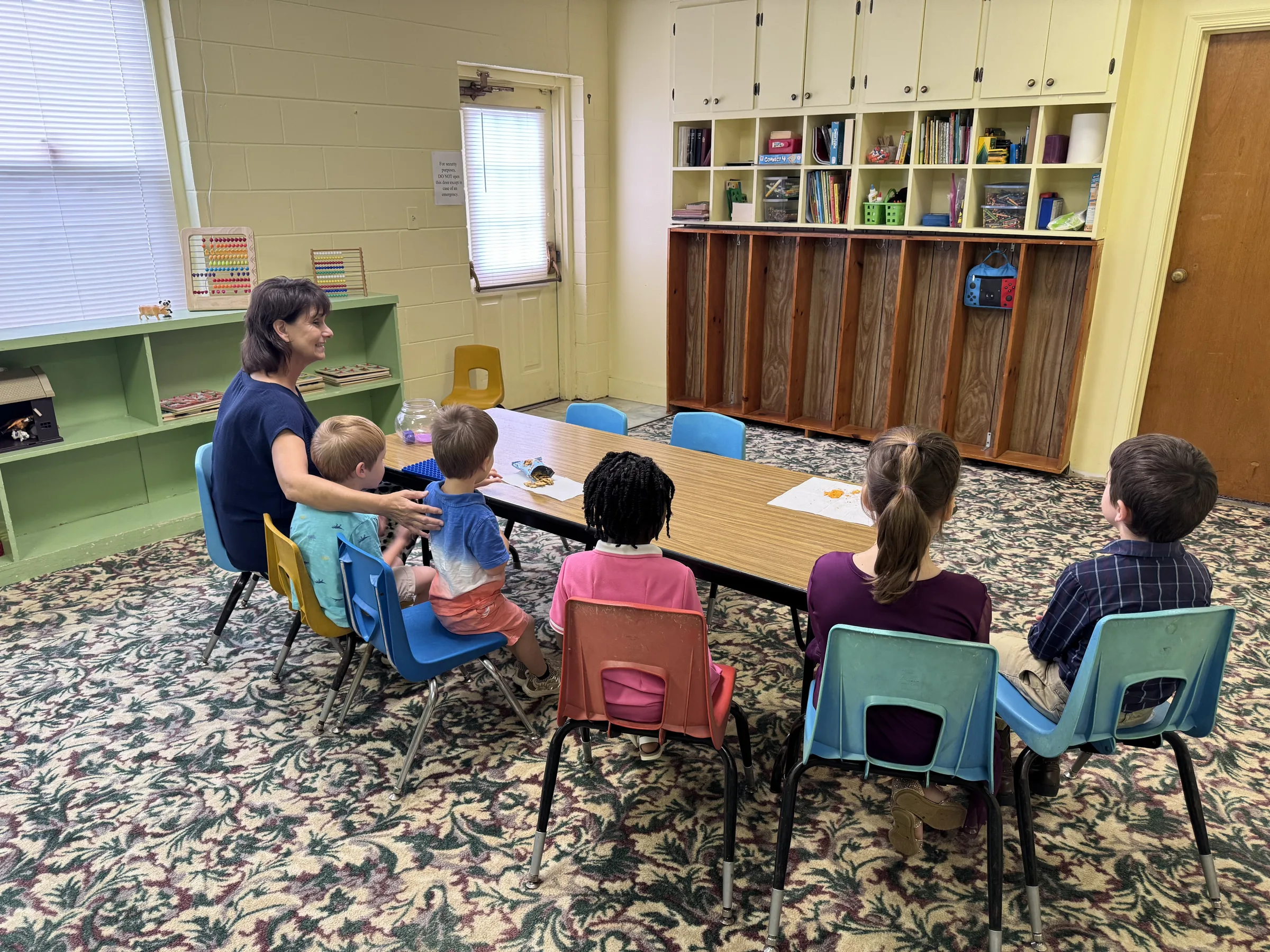 A teacher sits with children gathered around a classroom table at Grace